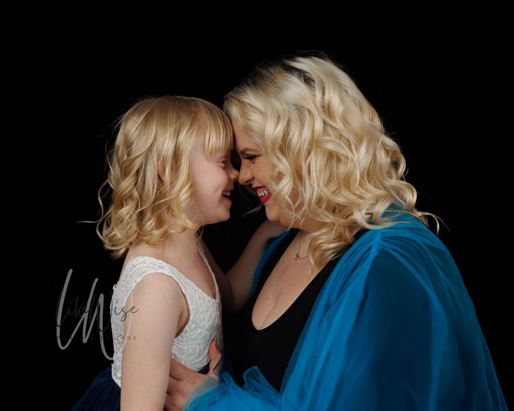 Two people with blonde curly hair share a tender moment against a dark background while wearing white and blue clothing.