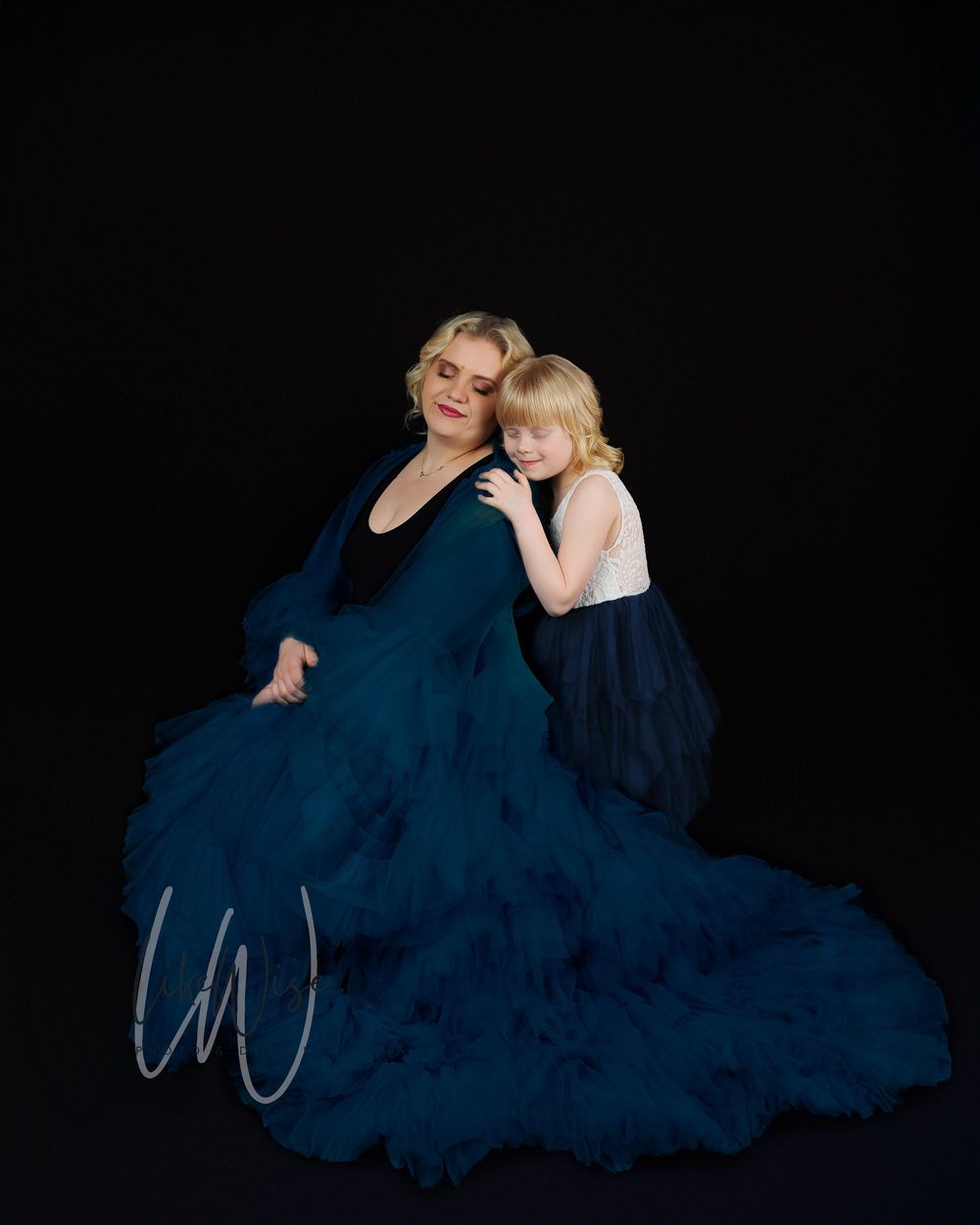 Two people in coordinating navy blue dresses pose together against a dark backdrop in an elegant studio portrait.