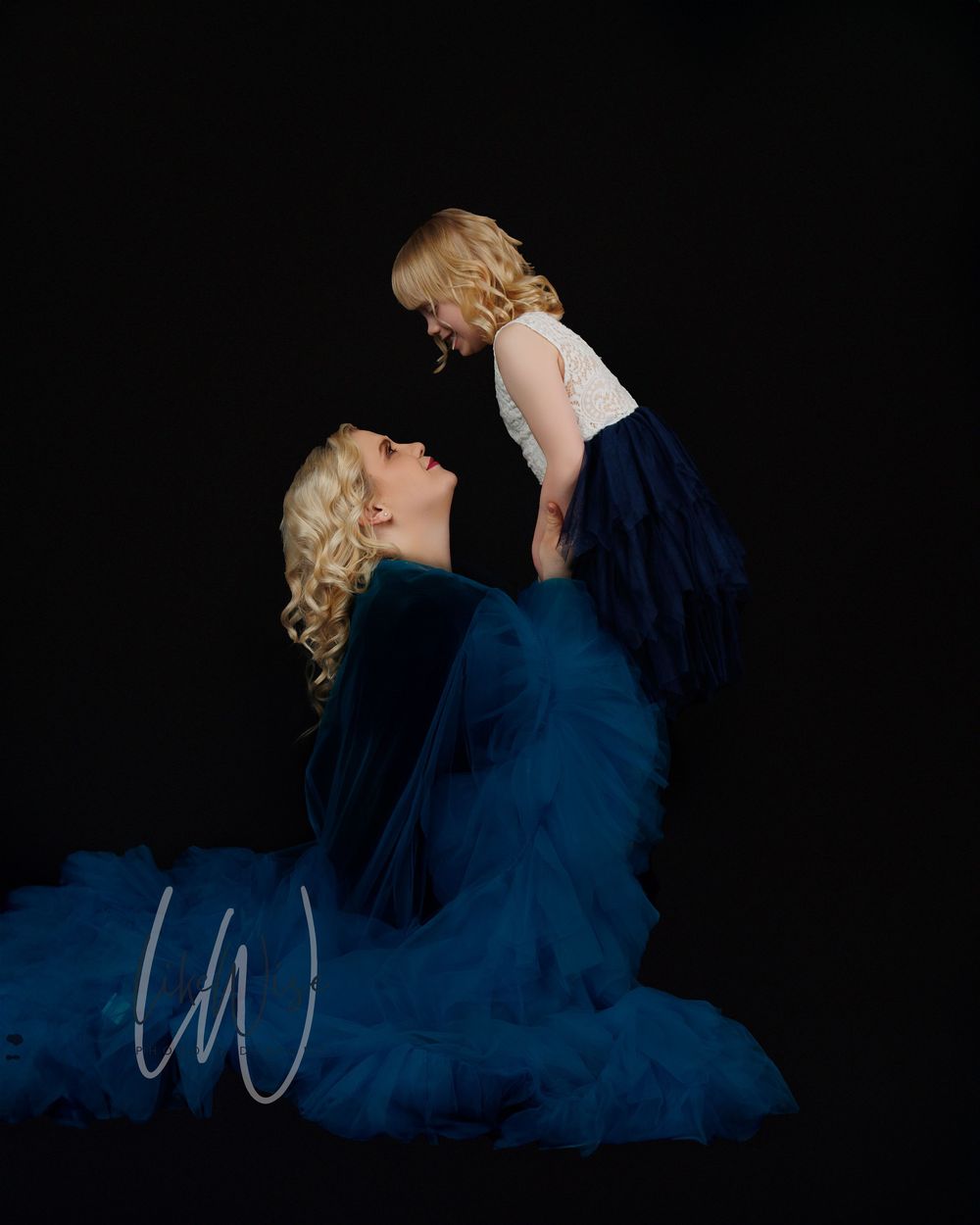 Mother and child wearing matching navy blue dresses pose for an artistic portrait against a black background.