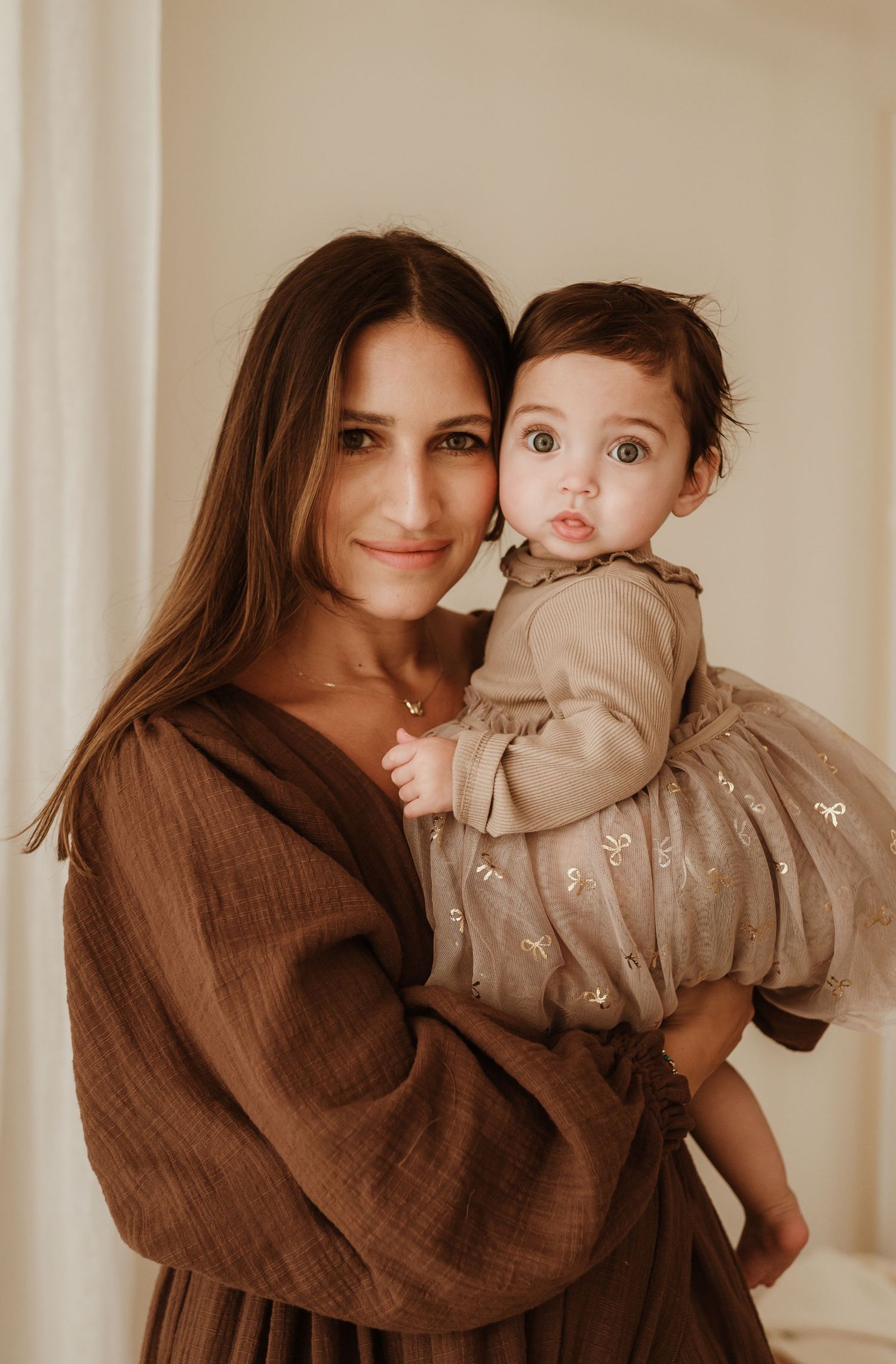 A woman in a brown dress holds a baby wearing a beige dress against a neutral background.