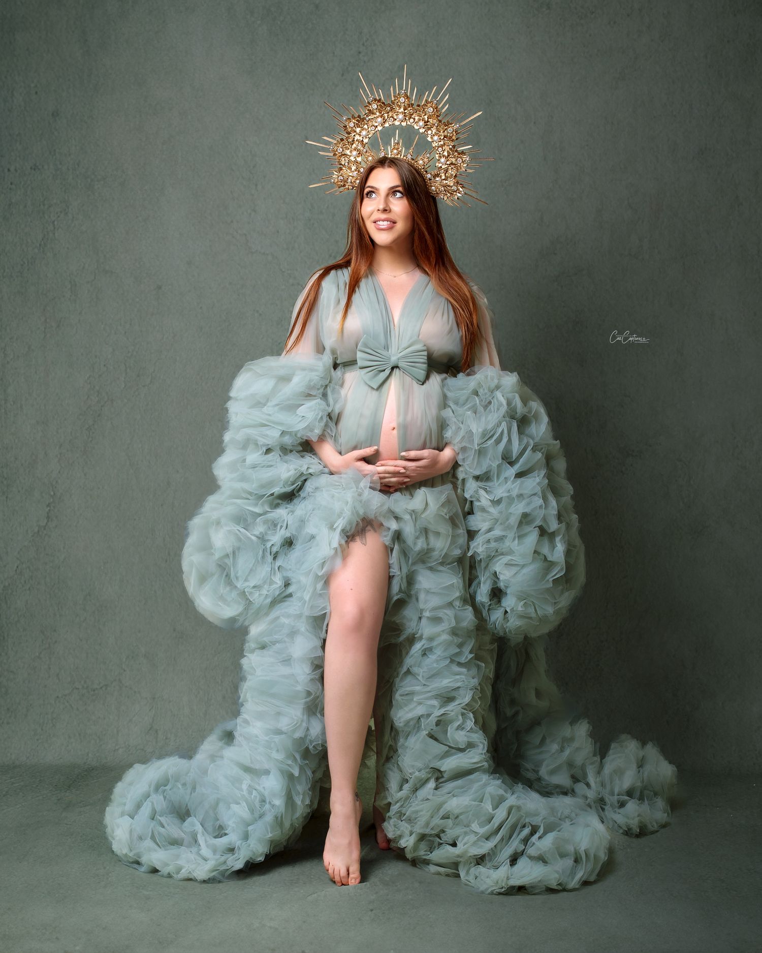 Dreamy studio portrait: woman in a  sage green  flowing maternity gown and gold crown against green backdrop.