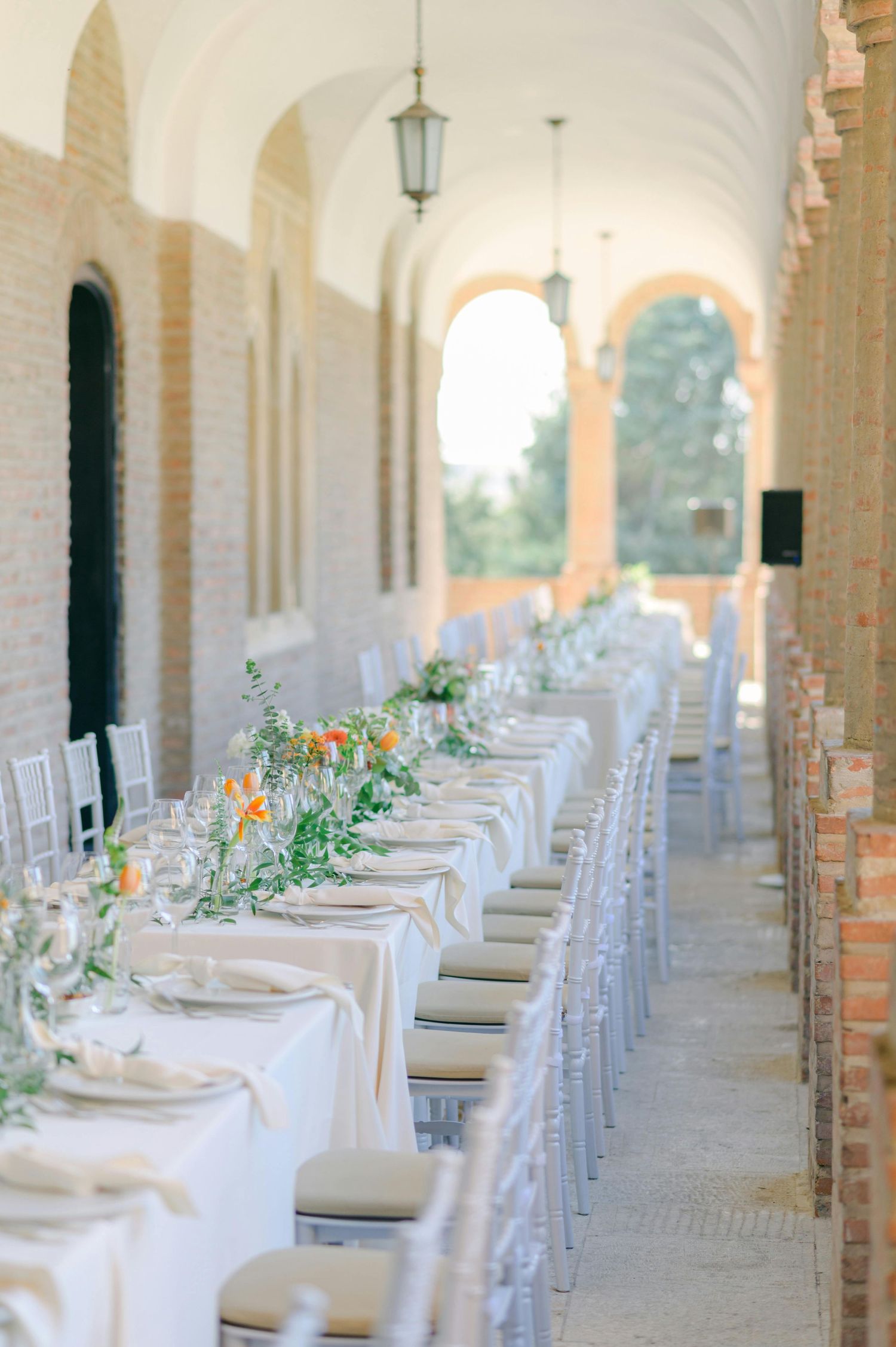 Elegant table setting along arched corridor with white linens and floral centerpieces.