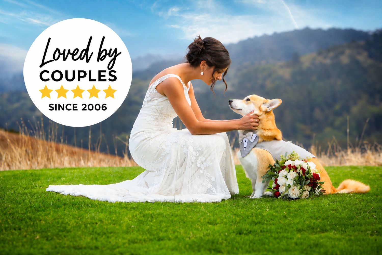 A bride in a white dress shares a tender moment with her dog on a grassy hilltop with mountains in the background.
