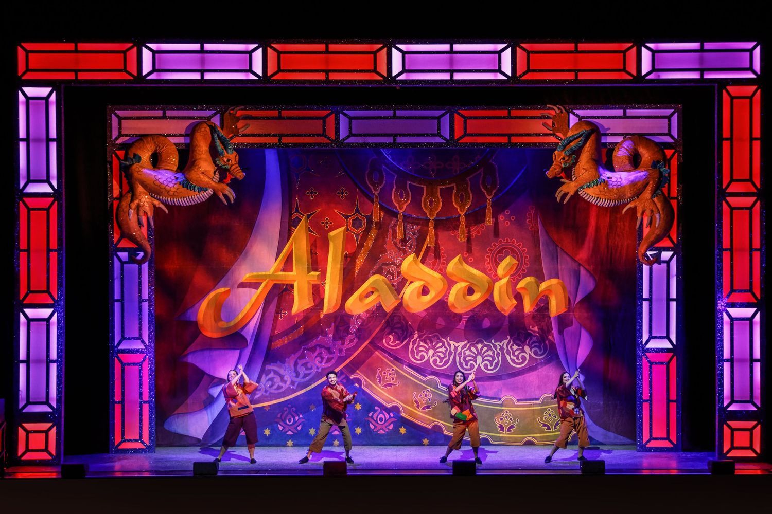 Aladdin musical theater stage with red and purple border design featuring performers in traditional costumes.