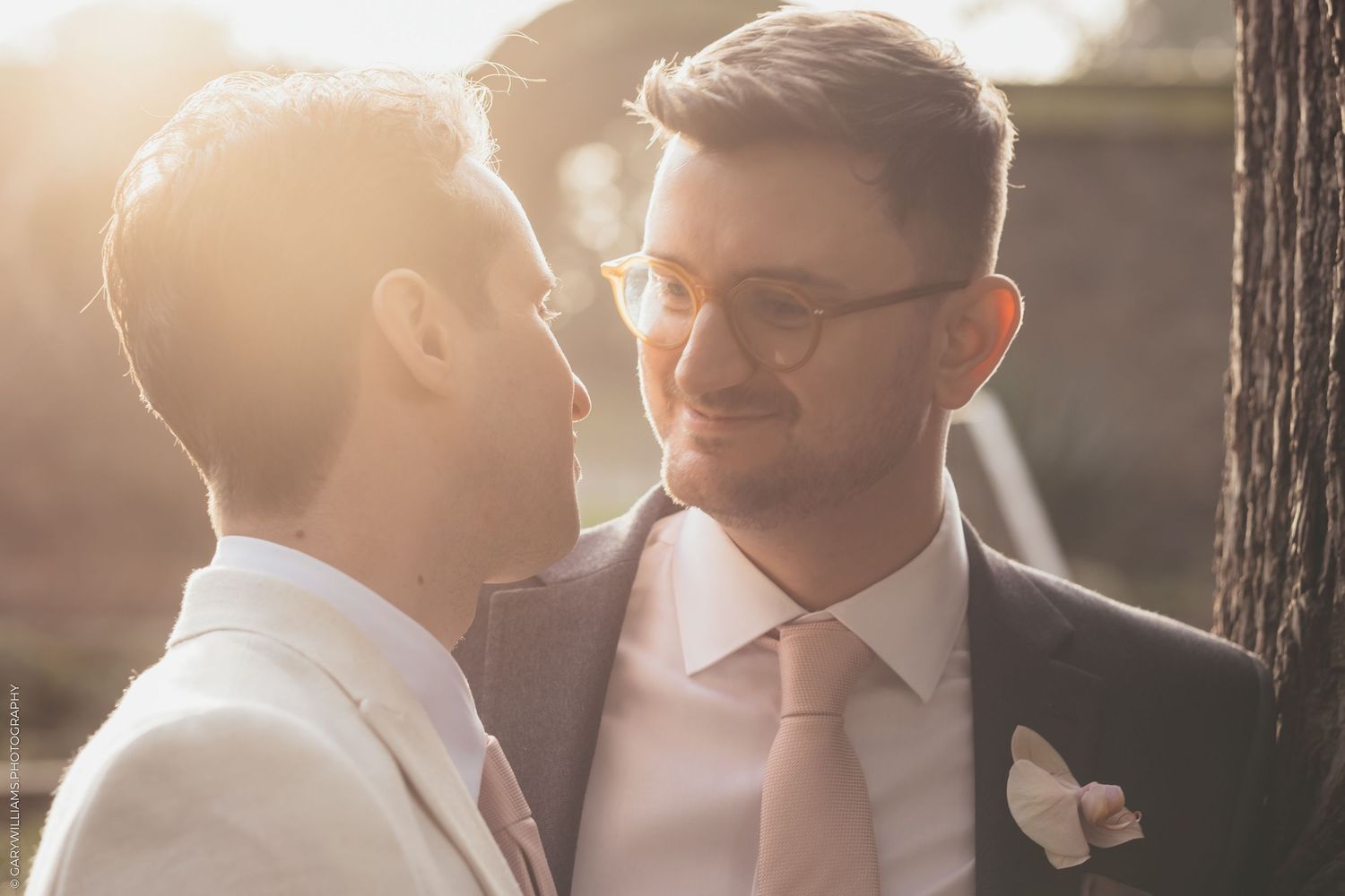 Two grooms in formal wedding attire share an intimate moment in warm sunlight wearing suits and ties.