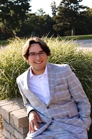 A person in a gray checkered blazer sits on a stone wall near ornamental grasses.