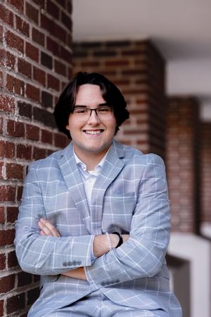 Indoor portrait against a brick wall wearing a light blue checkered blazer.