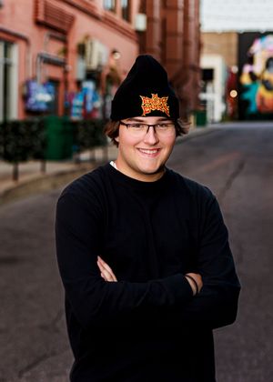 Person in black sweater poses on urban street with string lights overhead.