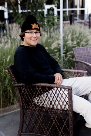 Person wearing black sitting in a brown wicker chair outdoors with greenery in background.