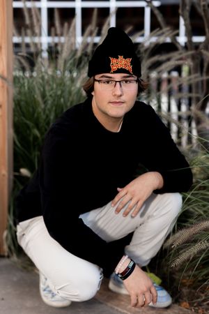 Person in black clothing and winter hat crouching near decorative grasses.