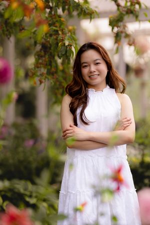 Portrait in a white sleeveless dress standing among blooming flowers in a garden setting.