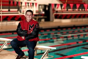 Athlete sits on starting block wearing varsity jacket at indoor swimming pool.