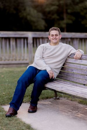 Someone in a beige sweater and blue jeans sits casually on a wooden bench outdoors.