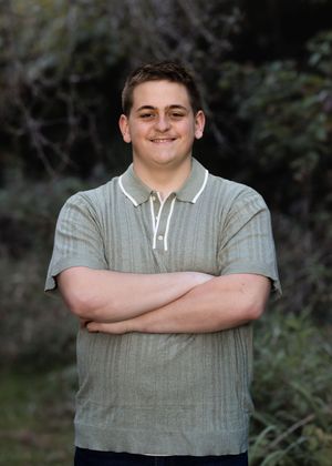 A person wearing a gray polo shirt poses with arms crossed against a natural backdrop.