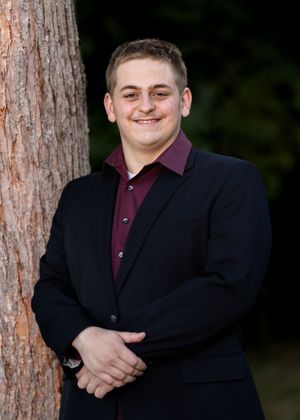 Someone wearing a black blazer and burgundy shirt poses between trees in an outdoor setting.