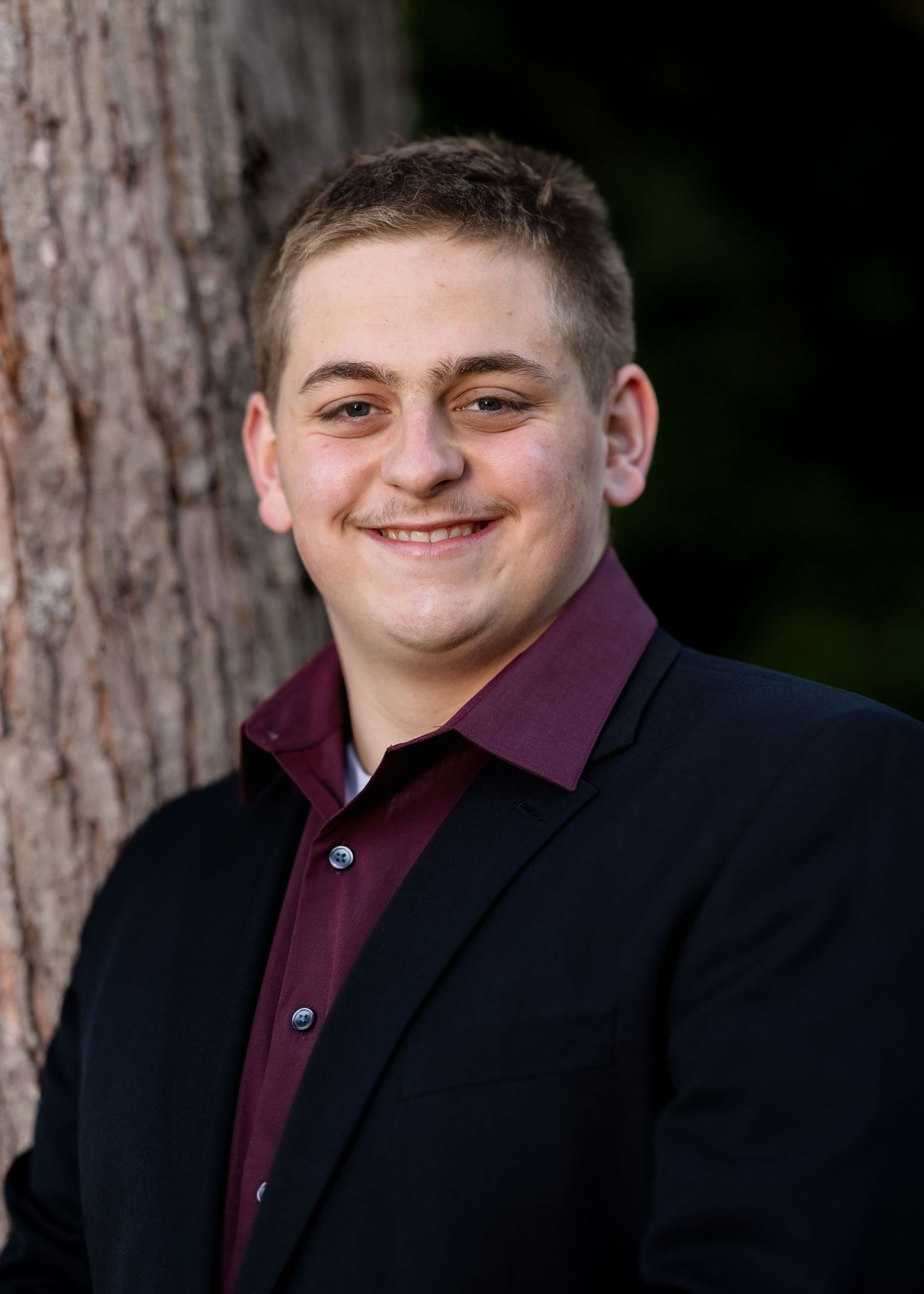 A person in formal attire stands against a backdrop of pine trees during a photo session.