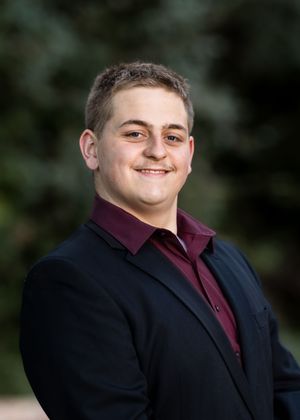 Someone wearing a black suit jacket and maroon shirt poses for formal portraits outdoors.
