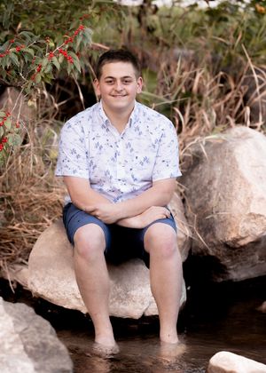 Someone wearing a white floral shirt and blue shorts sits casually on large rocks by a stream in nature.