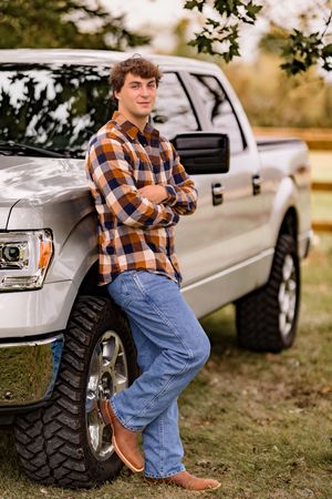 A person in orange plaid shirt and jeans poses against a silver pickup truck with large off-road tires.
