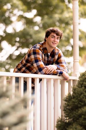 A person in a plaid flannel shirt leans casually on a white porch railing during an outdoor photoshoot.