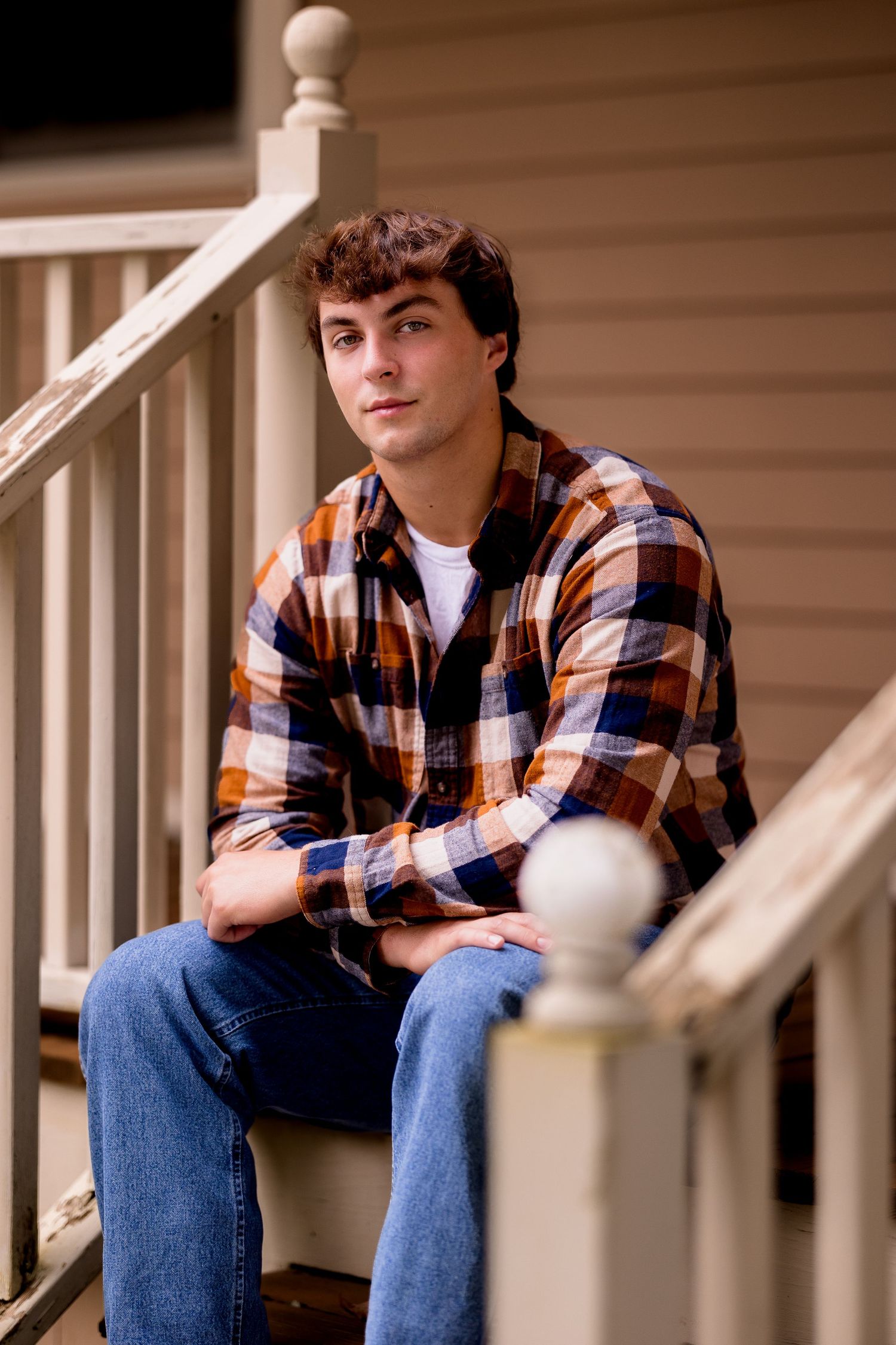 A person in plaid shirt and jeans sits casually on white porch steps during a photo session.