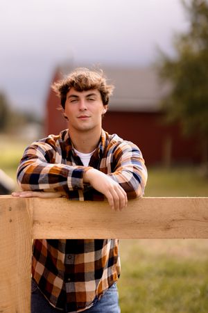 A person in a plaid shirt leans on a wooden fence post in a rural setting.