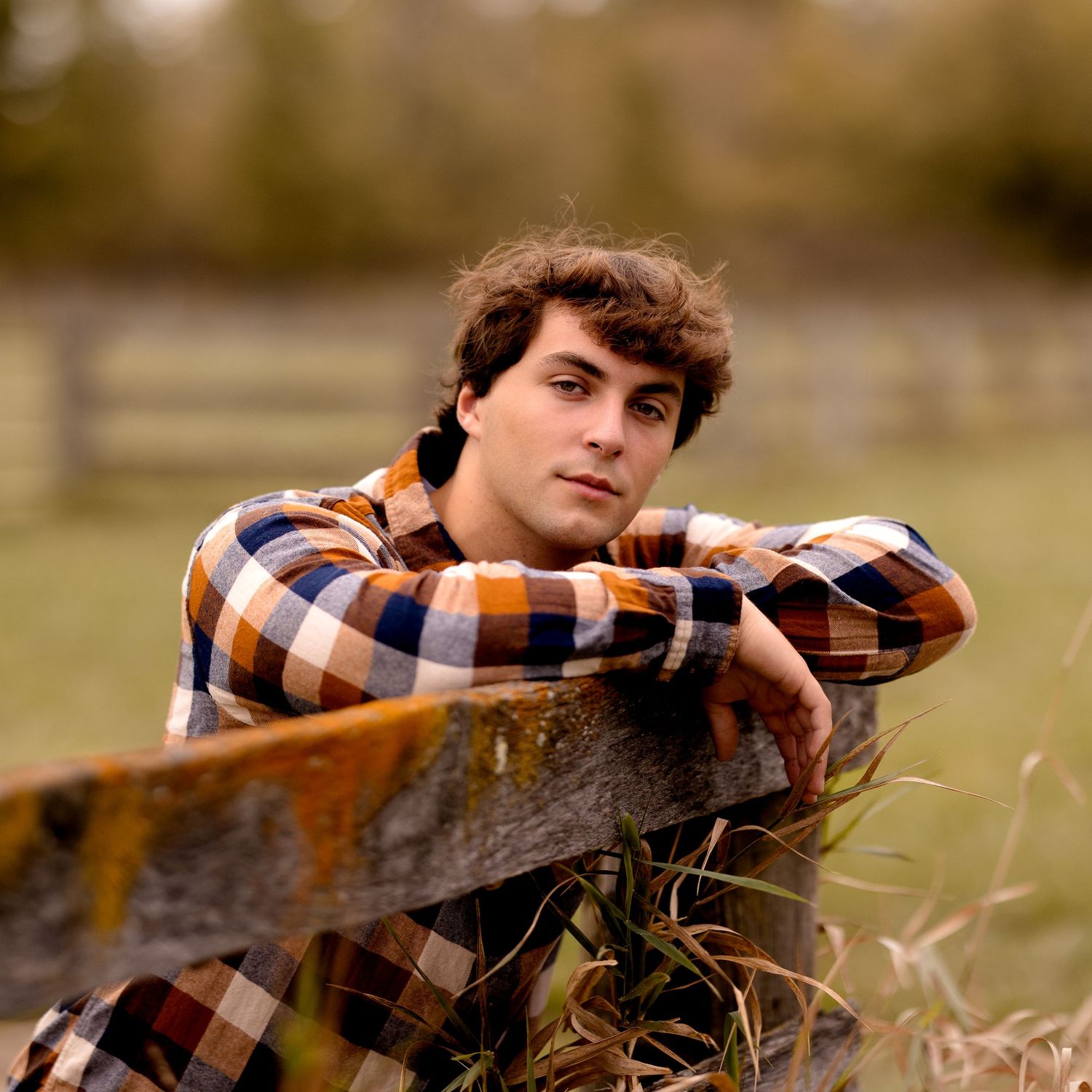Someone in an orange plaid shirt leans against a wooden fence in a rural setting.