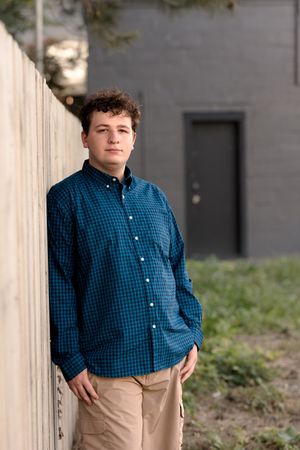 Person in blue checkered button-down shirt and khaki pants standing near concrete wall.