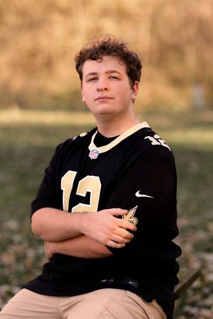 A person in a black Saints jersey stands with crossed arms against a blurred natural background.