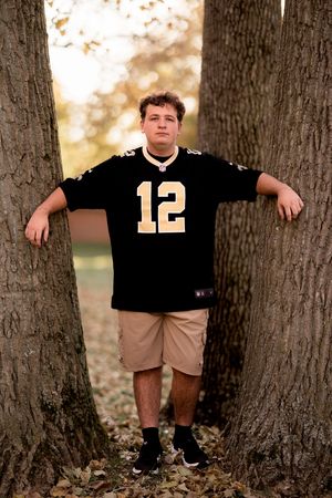 A person in a Saints jersey and khaki shorts poses between large tree trunks covered in fall leaves.