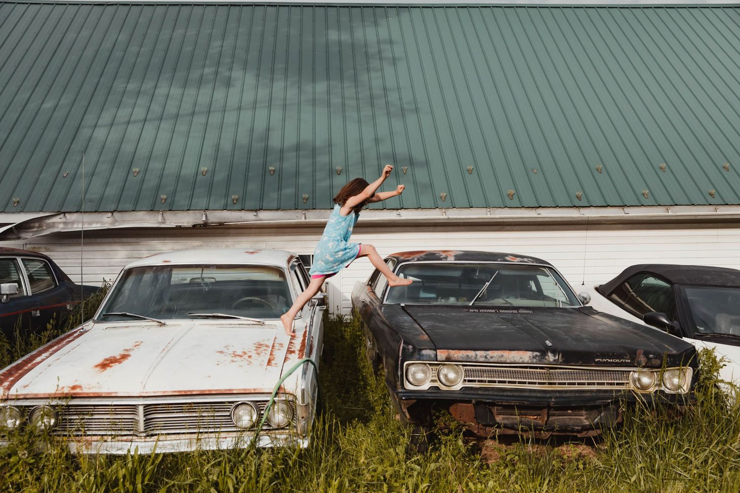 Image of a girl jumping across old cars during a documentary style family photo shoot at the families home