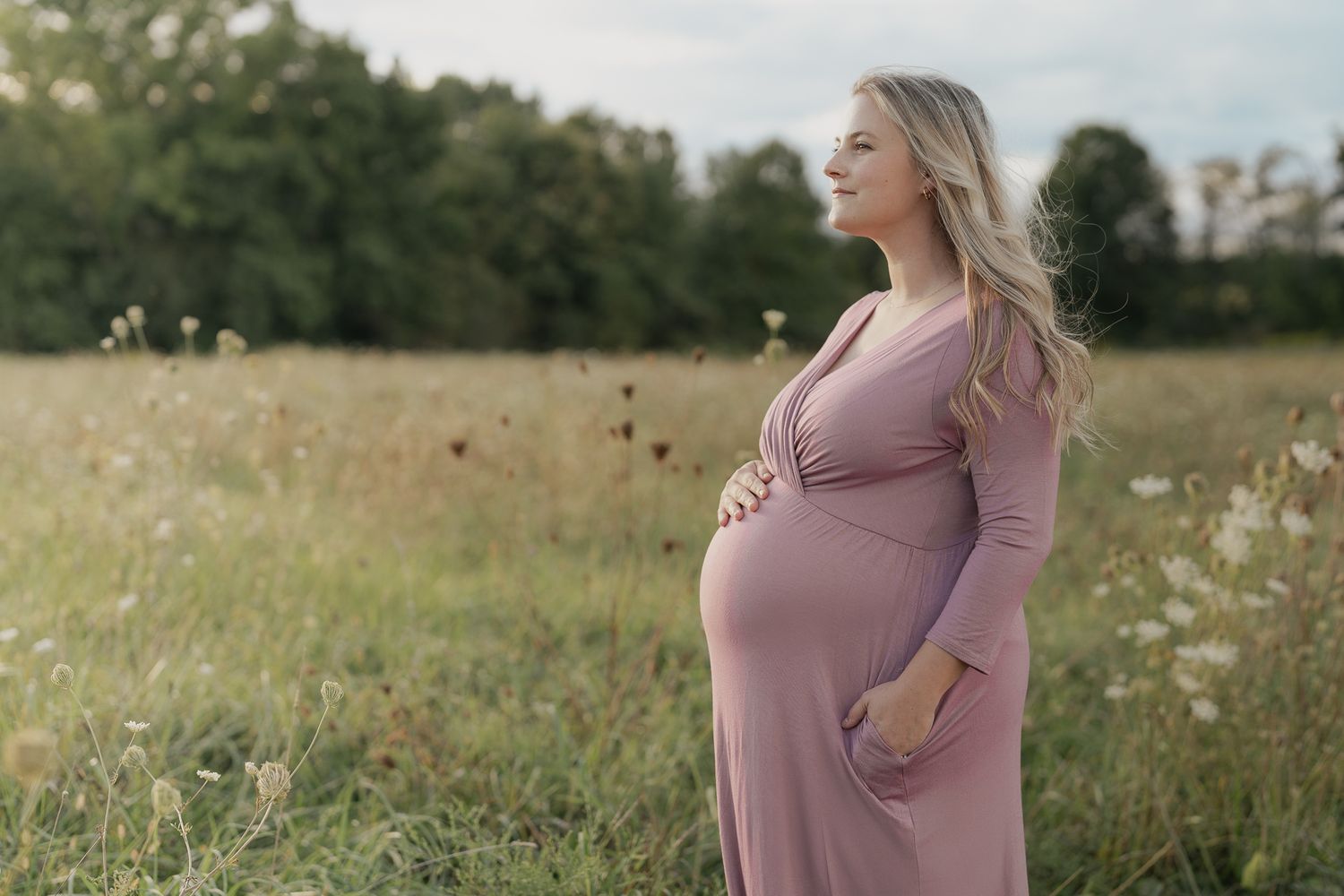 A person in a mauve dress stands in a sunlit meadow with wildflowers during sunset.