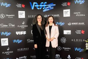 Two women pose together at a red carpet event with blue and white logos visible on the backdrop.