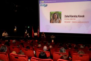 A dimly lit theater with red seats shows a presentation screen during an event or conference.