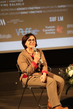 A speaker wearing glasses and a beige outfit sits on stage with a microphone during a presentation event.