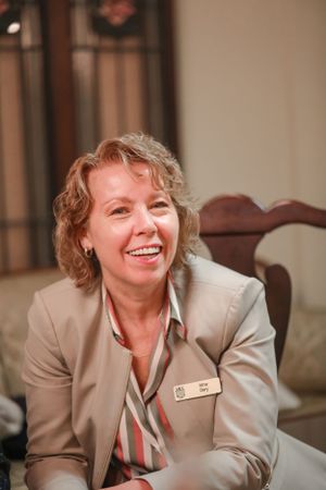 A professional wearing a beige blazer and striped shirt smiles warmly while sitting in an office setting.