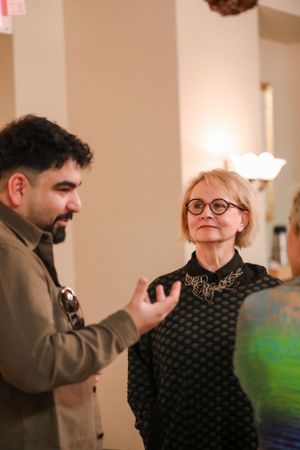 Two people engaged in conversation at an indoor event, with one wearing a brown jacket and another in a black patterned top.