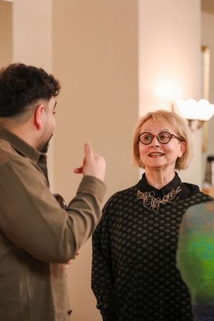 Two people engaged in an animated conversation, one wearing a black polka dot sweater and glasses, the other gesturing.