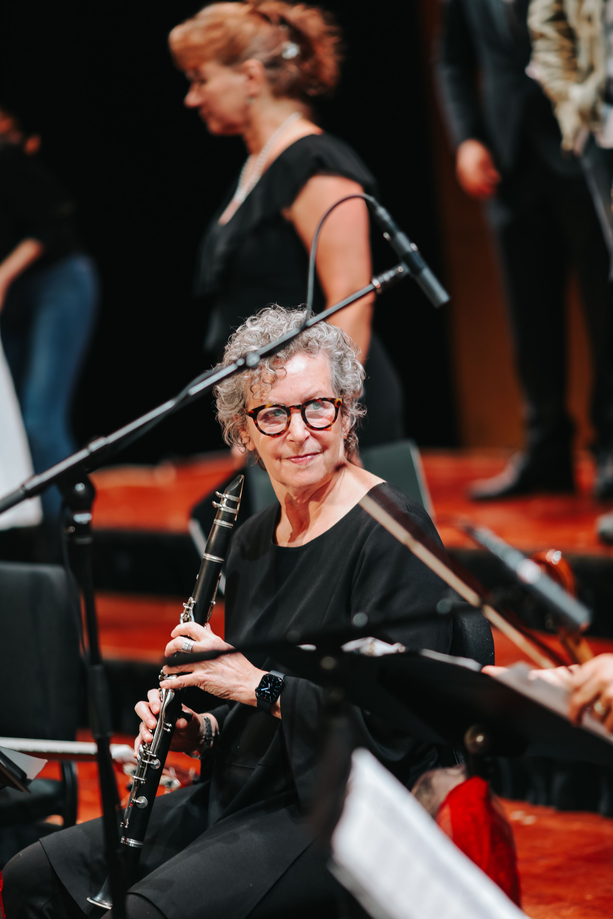 Senior musician in black attire smiles while holding a clarinet during an orchestra performance on red carpet.