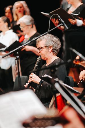 Musician in black attire plays clarinet with choir and orchestra performing in background on dark stage.