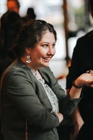 Smiling person in a grey blazer and white lace top wearing dangling earrings looking to the side during a social event.