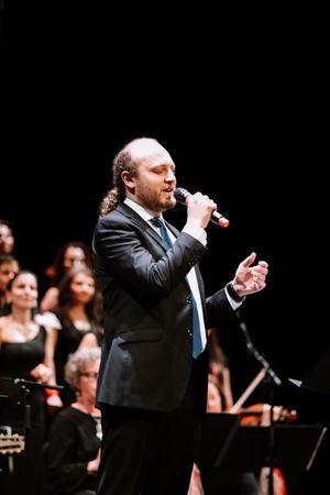 A performer in a black suit and blue tie sings into a microphone on stage with a choir visible in the background.
