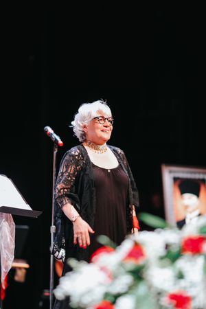 An older performer in a black lace dress and pearls stands on stage with red and white flowers in the foreground.