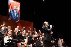 A concert performance featuring a large choir and orchestra on stage with banners displayed above.