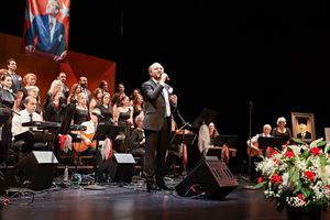 Orchestra performs on stage with conductor leading ensemble at formal concert venue with red floral decorations.