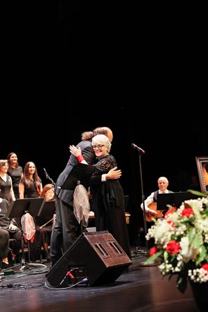 Two performers share an emotional embrace on a dark stage with flowers and musical equipment visible in the performance space.