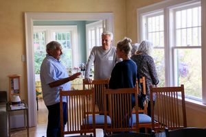 A group of people gather in a sunny room with large windows and wooden chairs for a casual meeting.