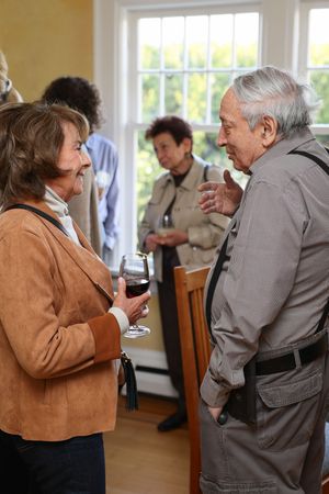 Two guests engage in conversation at a social gathering while holding wine glasses in a bright room with windows.