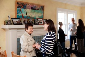 Two people chat over drinks at a cocktail party while others socialize in the background by a fireplace.