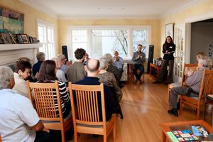 People gathered in a sunny living room for an intimate performance or presentation with wooden chairs arranged in rows.